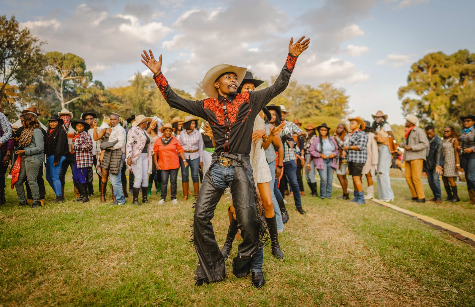 A man wearing chaps and a cowboy hat leads a line of dancing people at an outdoor music festival.
