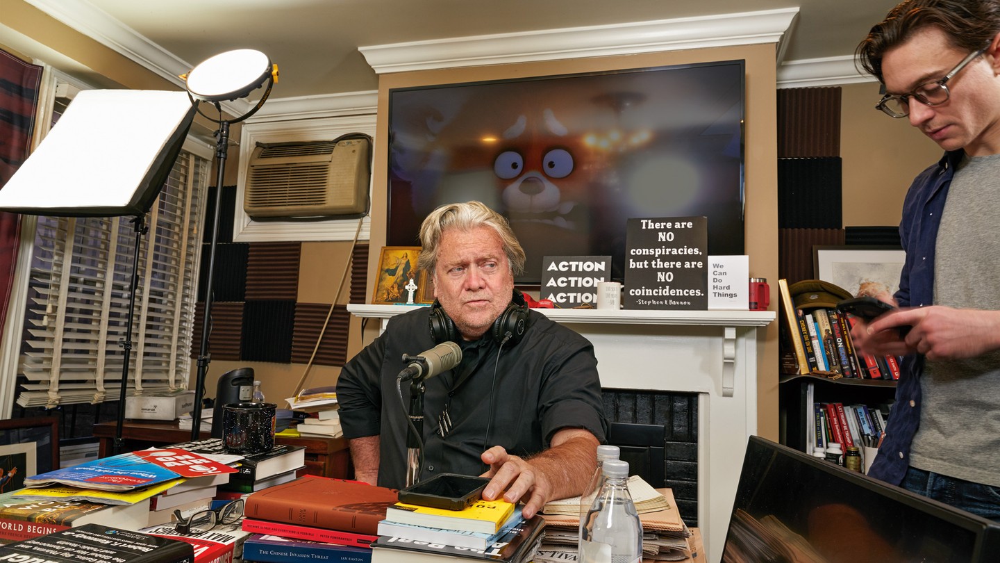 Photo of Steve Bannon sitting in cluttered home studio at microphone on desk covered with books near lighting equipment, in background a TV with still from movie "Turning Red" over fireplace mantel with signs