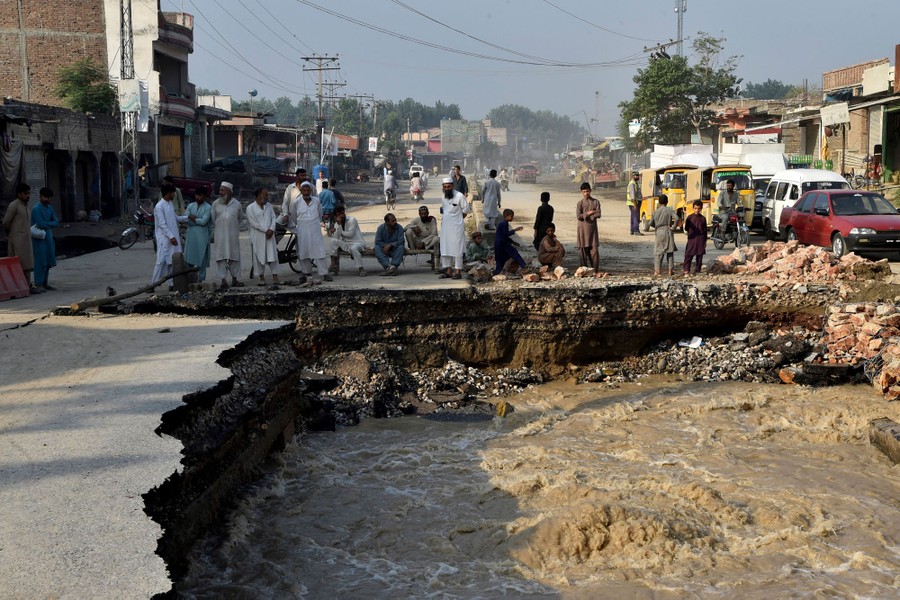 People stand on a heavily damaged road, with floodwater roiling in a wide hole.