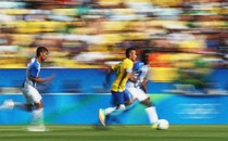 Three soccer players run toward the right side of the frame; the one in the center wears the distinctive yellow jersey of the Brazilian men's national team.