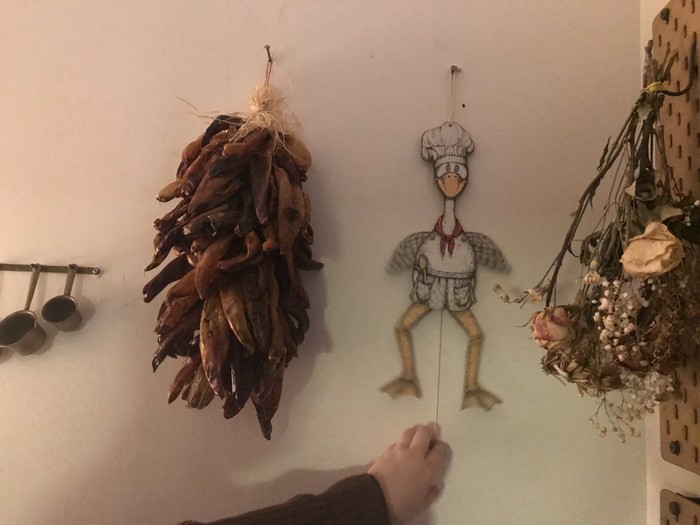 The wall of an apartment kitchen, decorated with a bundle of dried chile peppers and a wooden goose.