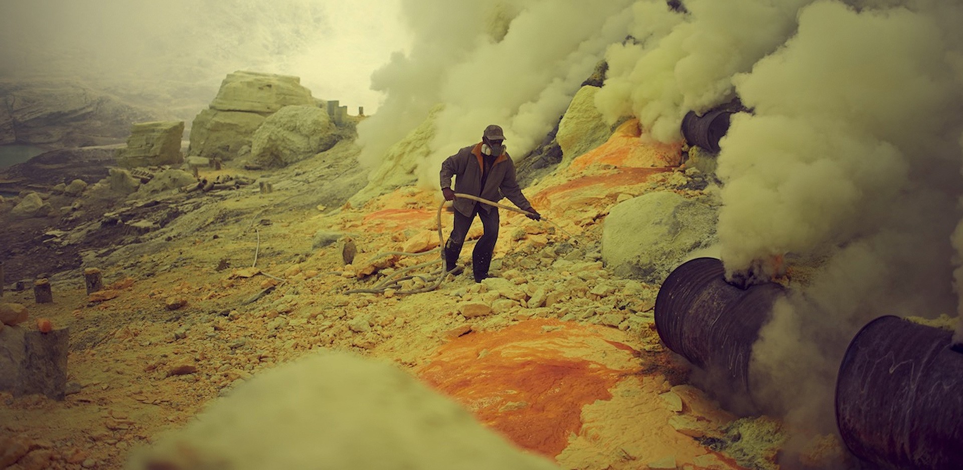 A man mining sulfur in Indonesia. There are plumes of smoke on the left, and the ground is covered with orange and grey rocks.