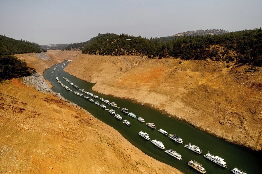 Two lines of boats sit anchored in a narrow channel of a mostly empty reservoir.