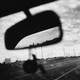 a black and white photo of the road and the cloudy sky seen out the front window of a car, the mirror taking up the front of the frame