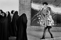 a woman in a headscarf walks in front of a mural showing Ruhollah Khomeini and toward a group of women in black chadors
