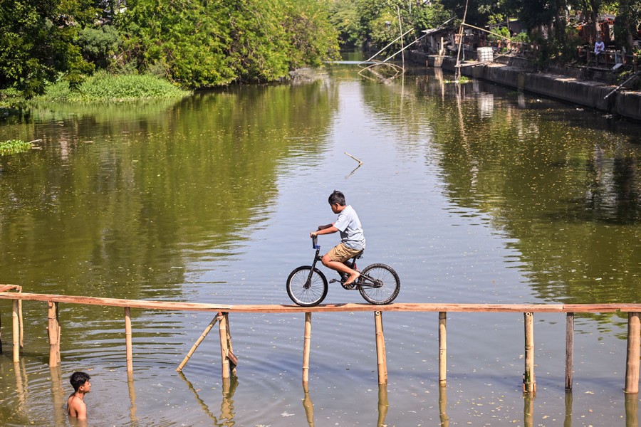 A child rides a bicycle on a narrow plank bridge over a calm river.