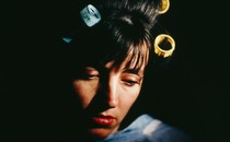 A woman with shadows on her face and curlers in her dark hair looks down, sadly.