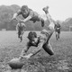 A black and white photograph of a rugby game in action, two players diving for the ball and three running behind them in striped uniforms in the distance on the field