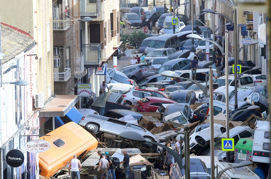 People stand next to dozens of piled-up cars in a narrow street.