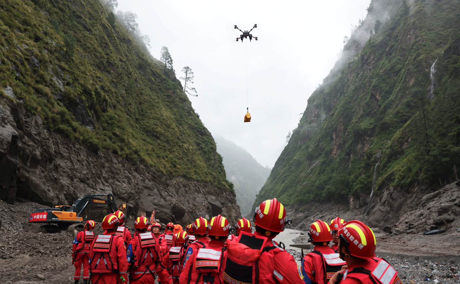 A group of rescue workers in a narrow mountain river valley look up toward a large drone carrying supplies on a long cable.