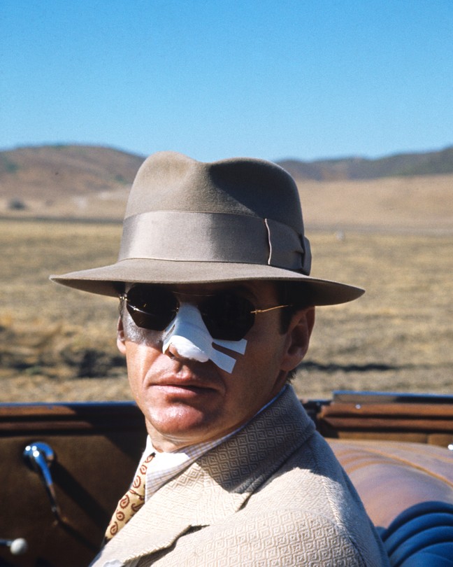 A movie still of Jack Nicholson, playing private eye J. J. Gittes in Chinatown (1974), in a convertible car with the top down, wearing a fedora and sunglasses with his nose covered in white medical tape.