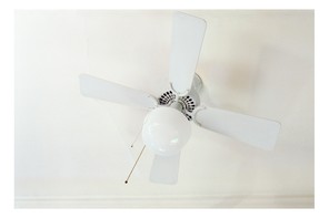 A gauzy photograph, closely cropped, of a white ceiling fan and an unlit bulb, on a white ceiling