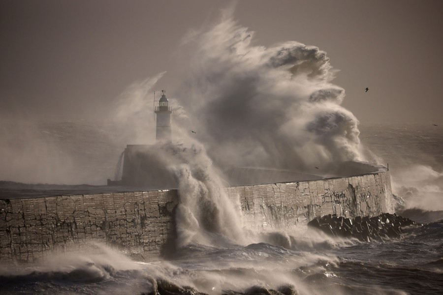 High waves crash into a harbor wall and lighthouse.