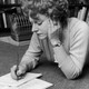 A photograph of Muriel Spark writing on a notebook on the floor