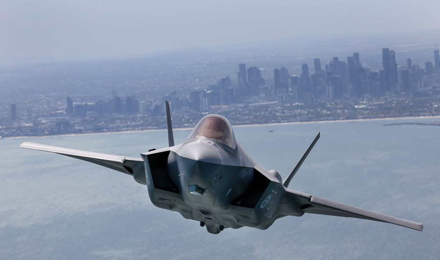 A fighter jet flies over a bay near a city.