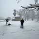 photo of person in winter gear and large camera on tripod covered with umbrella in snow-covered landscape with colonial house and split-rail fence