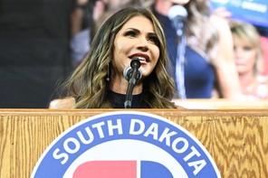 Kristi Noem stands at a podium at a 2023 rally in South Dakota and speaks into a microphone with a crowd behind her