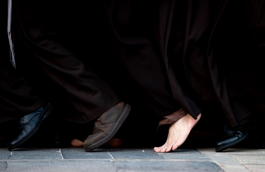 A close view of the feet of several people walking close together, wearing long robes. One of the people is barefoot.