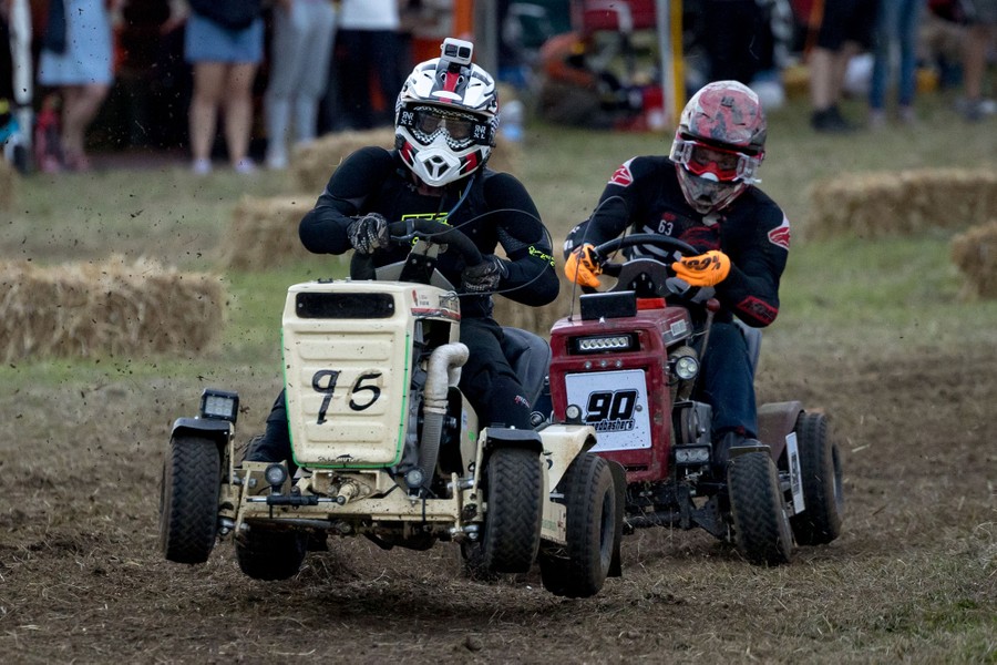 Two drivers wearing helmets race on a dirt track, on riding lawn mowers.