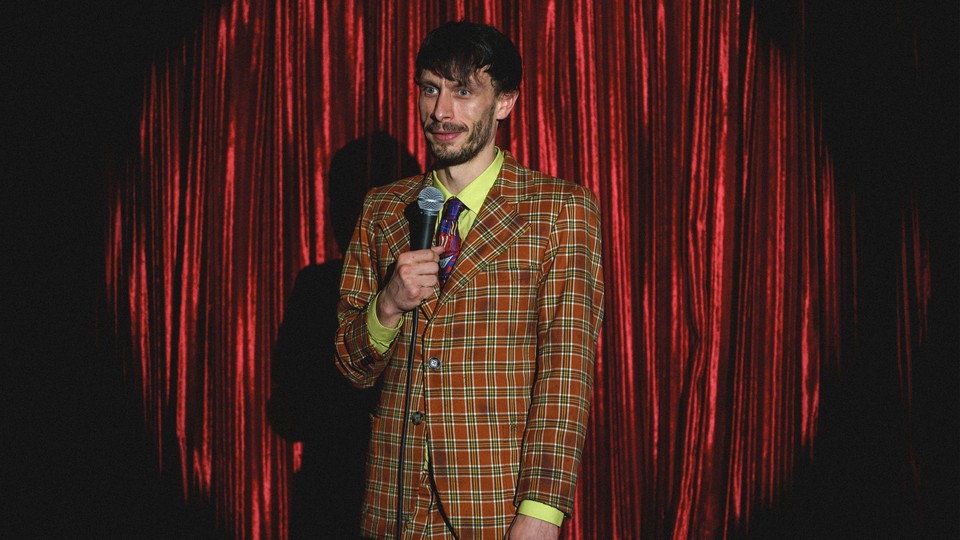 Richard Gadd in an orange plaid suit onstage, holding a microphone in front of red curtains