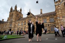 Two graduates toss their caps in the air while standing in front of a stately brick building.