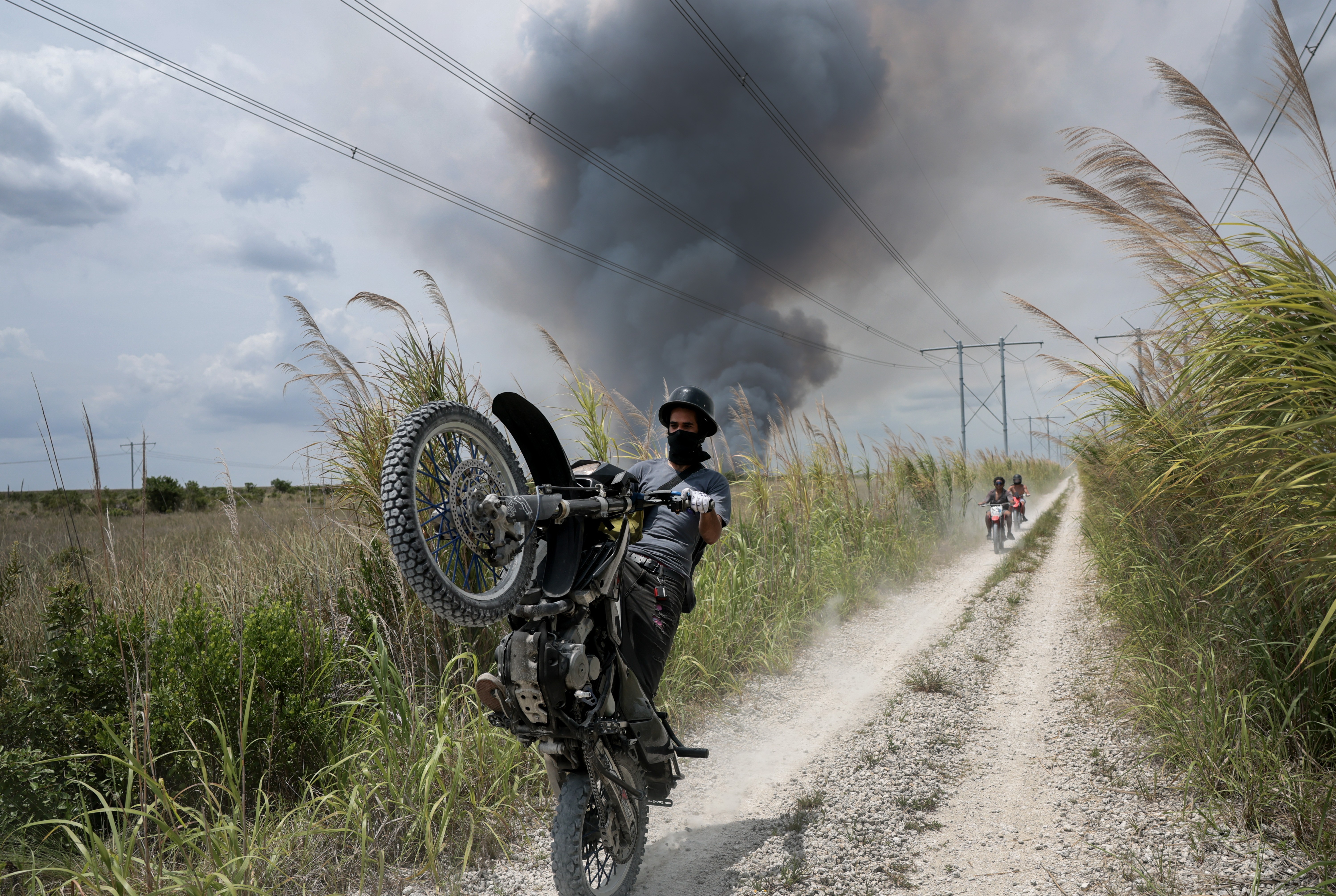 A motorcycle rider pops a wheelie in front of the photographer, while riding on a dirt road through grassland, with dark smoke rising in the background from a brushfire.