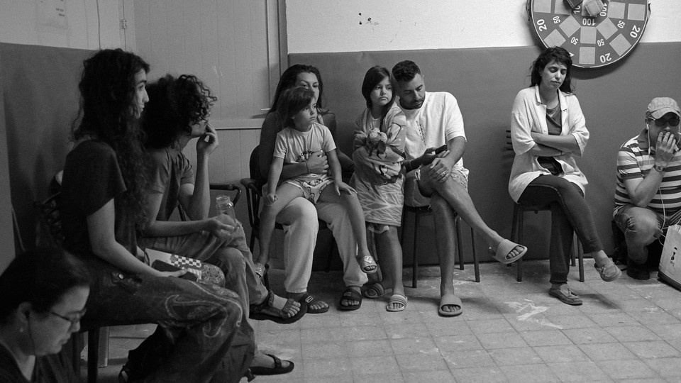An Israeli family and neighbors sit in chairs and on the floor in a Jerusalem bomb shelter.
