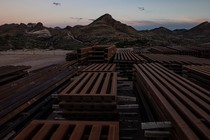 Stacks of steel bollards sit in a desert. In the background, a blasted mountain and Donald Trump's unfinished wall.
