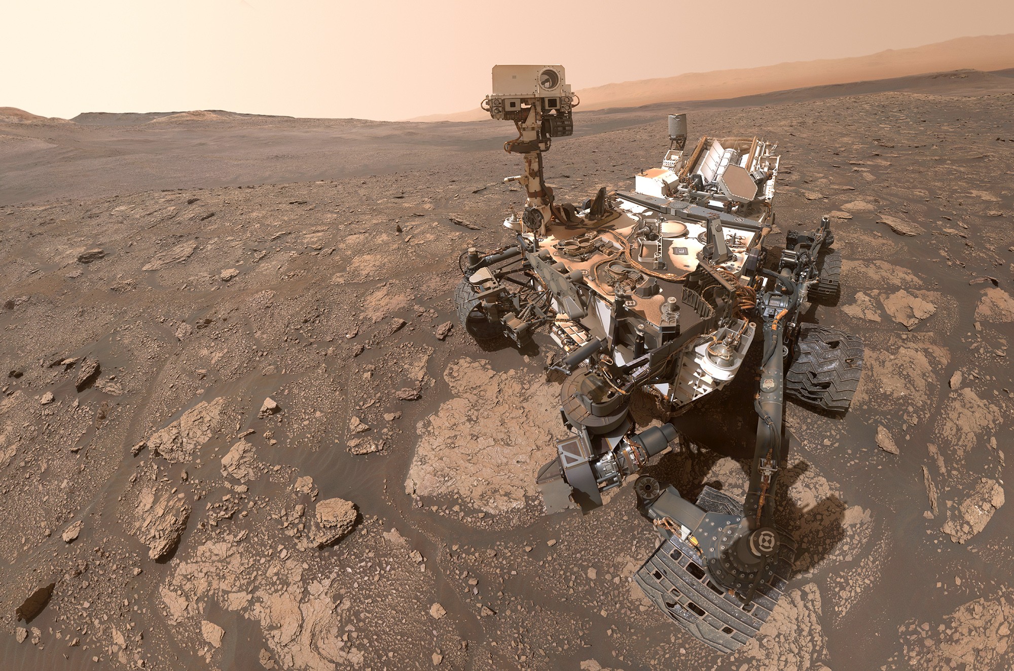 A selfie of a large Mars rover, with a reddish-colored dry, rocky, and dusty Martian landscape in the background.