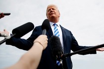 Donald Trump stands, arms outstretched, behind a row of hands holding microphones.