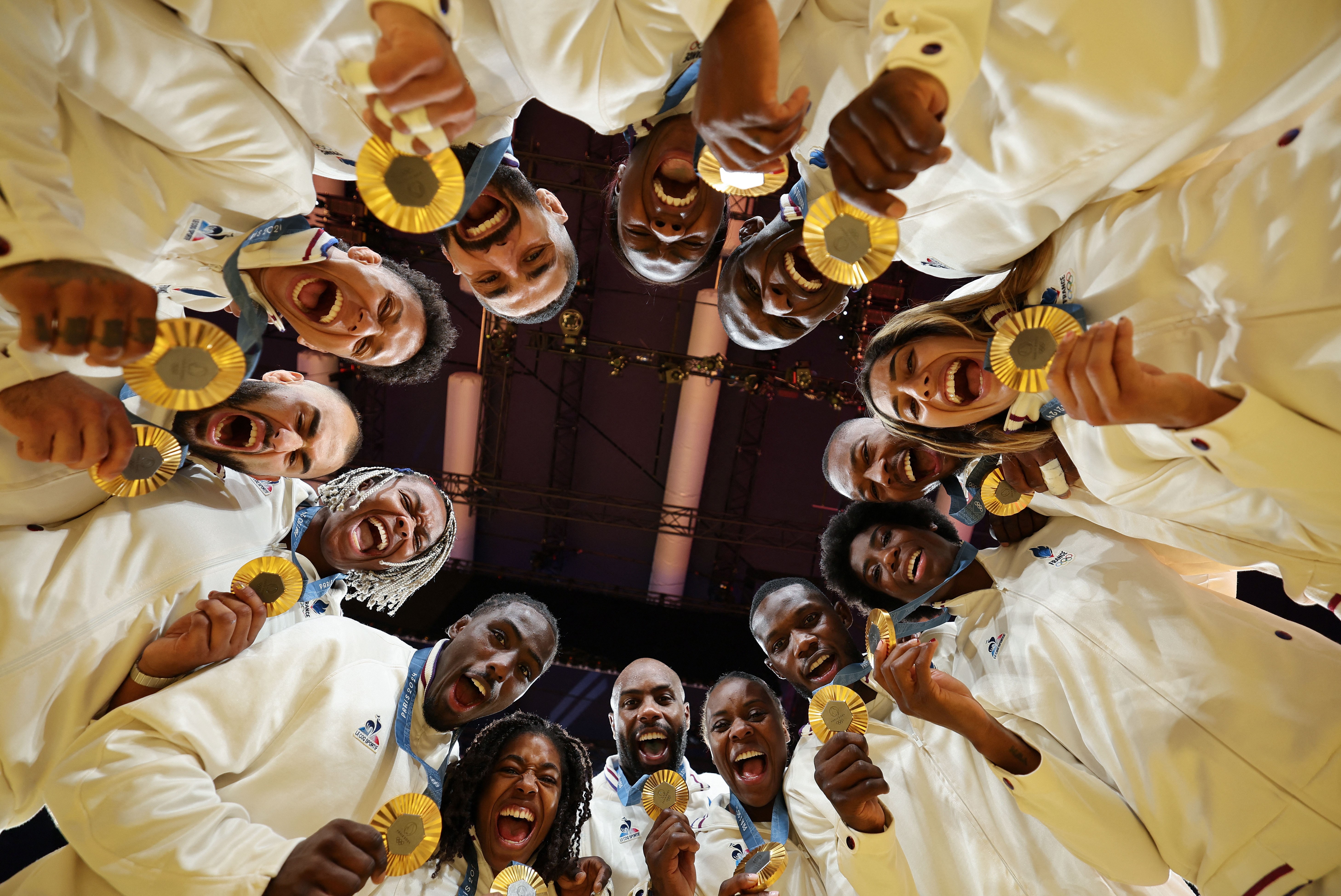 A view looking up at fourteen athletes, gathered in a circle, all holding gold medals, cheering and smiling down toward the camera.