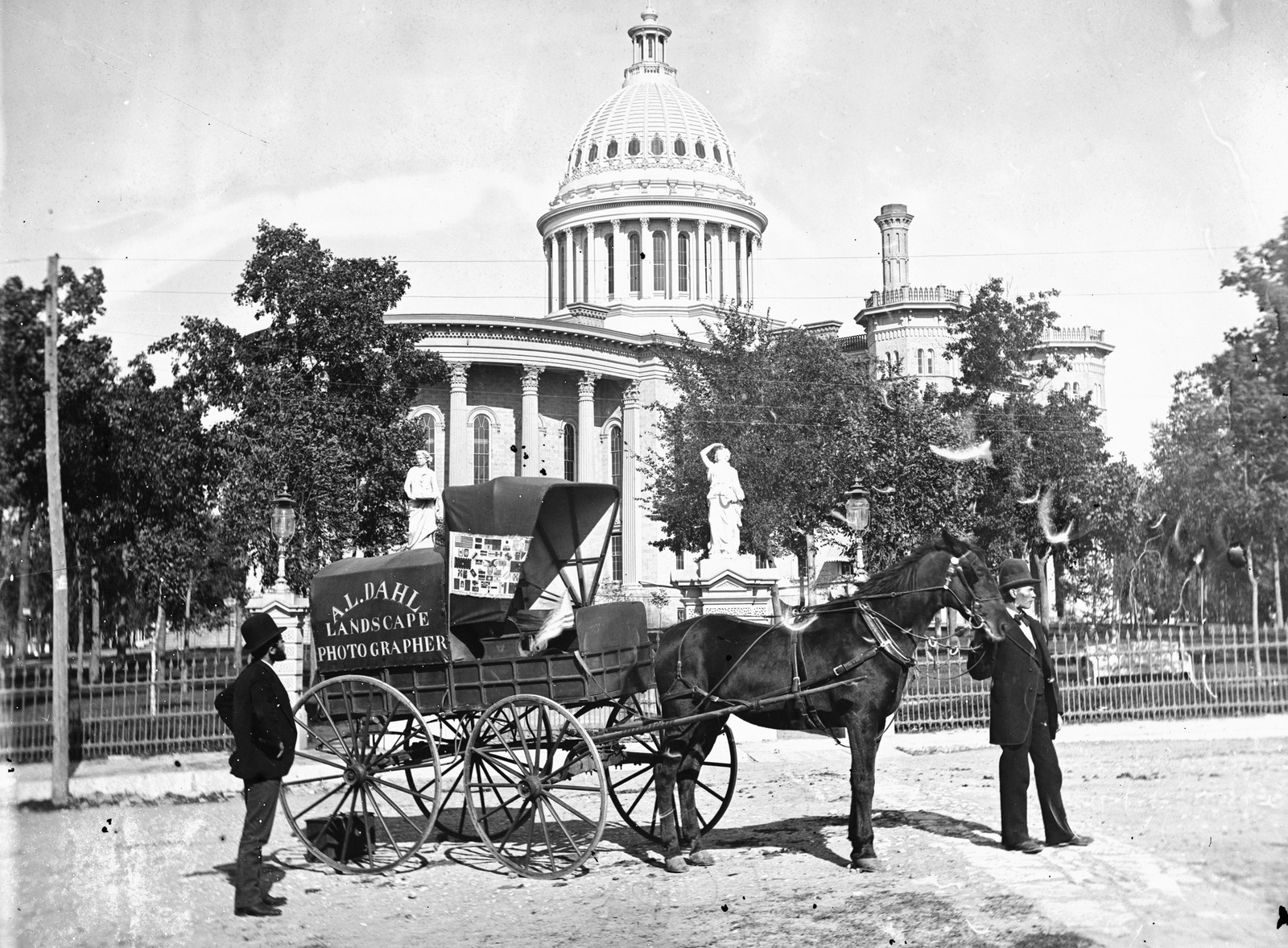 Two men pose beside a small horse-drawn cart in front of a state capitol building. The sign on the cart reads: 