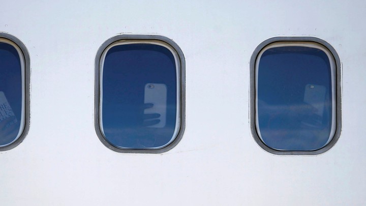 Two windows on the side of an airplane show hands holding up smartphones inside.