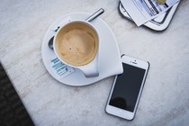 A smartphone sits unattended by a cup of coffee on a table.