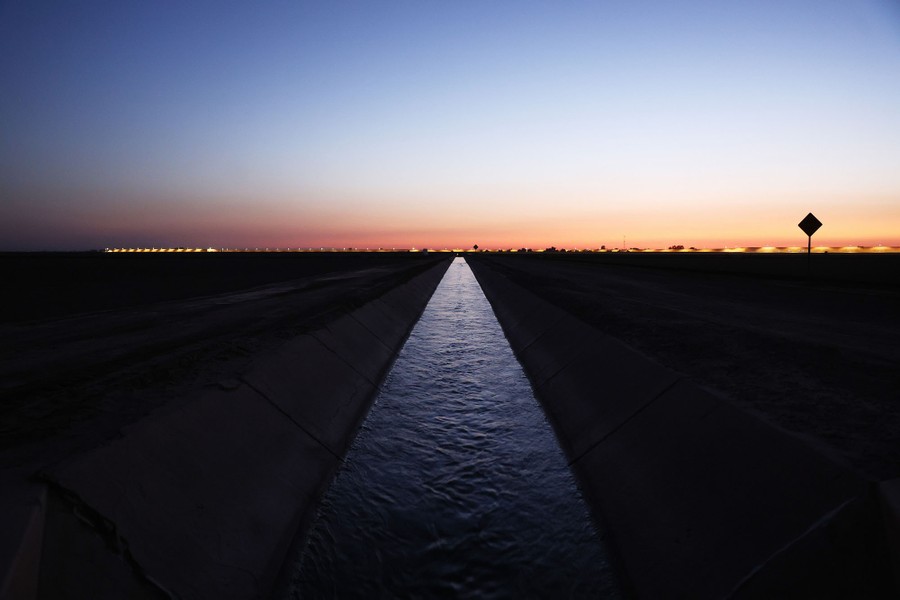 An aerial view of water flowing through a straight irrigation ditch in a desert, with the sun low in the sky