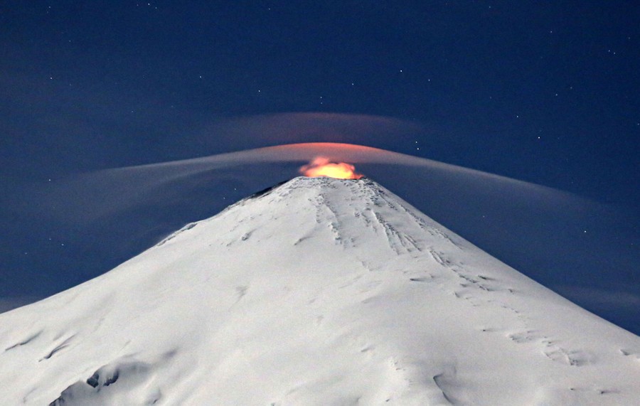 A glow emanates from the crater atop a snow-covered volcano.