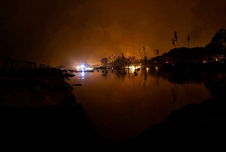 A night view of distant fires burning near buildings, illuminating the smoke that rises above.