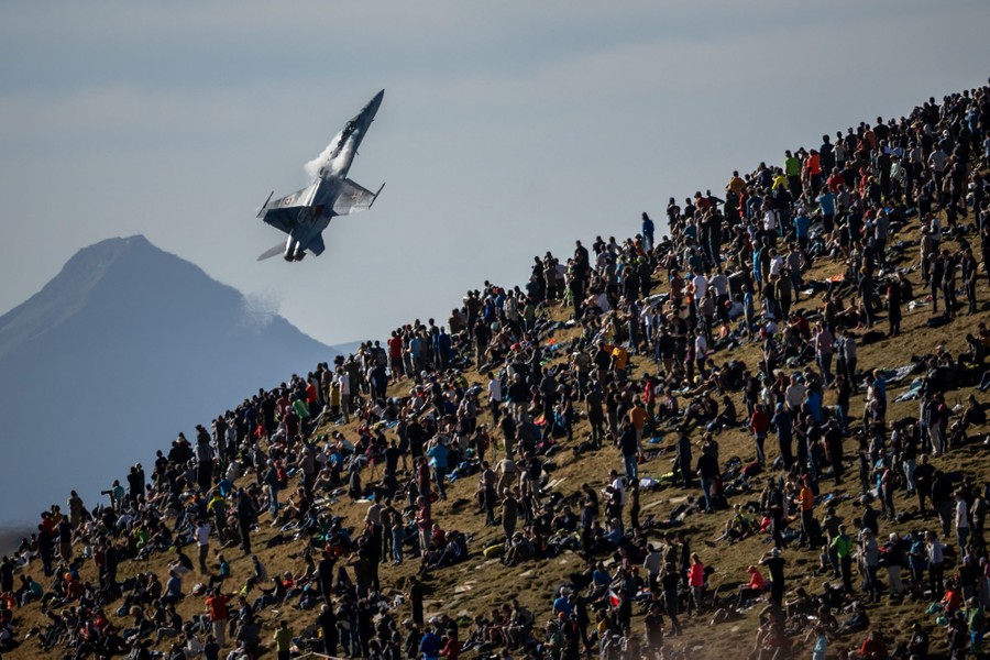 A steep, grassy hillside is covered with a crowd of spectators watching as a fighter jet flies close by.