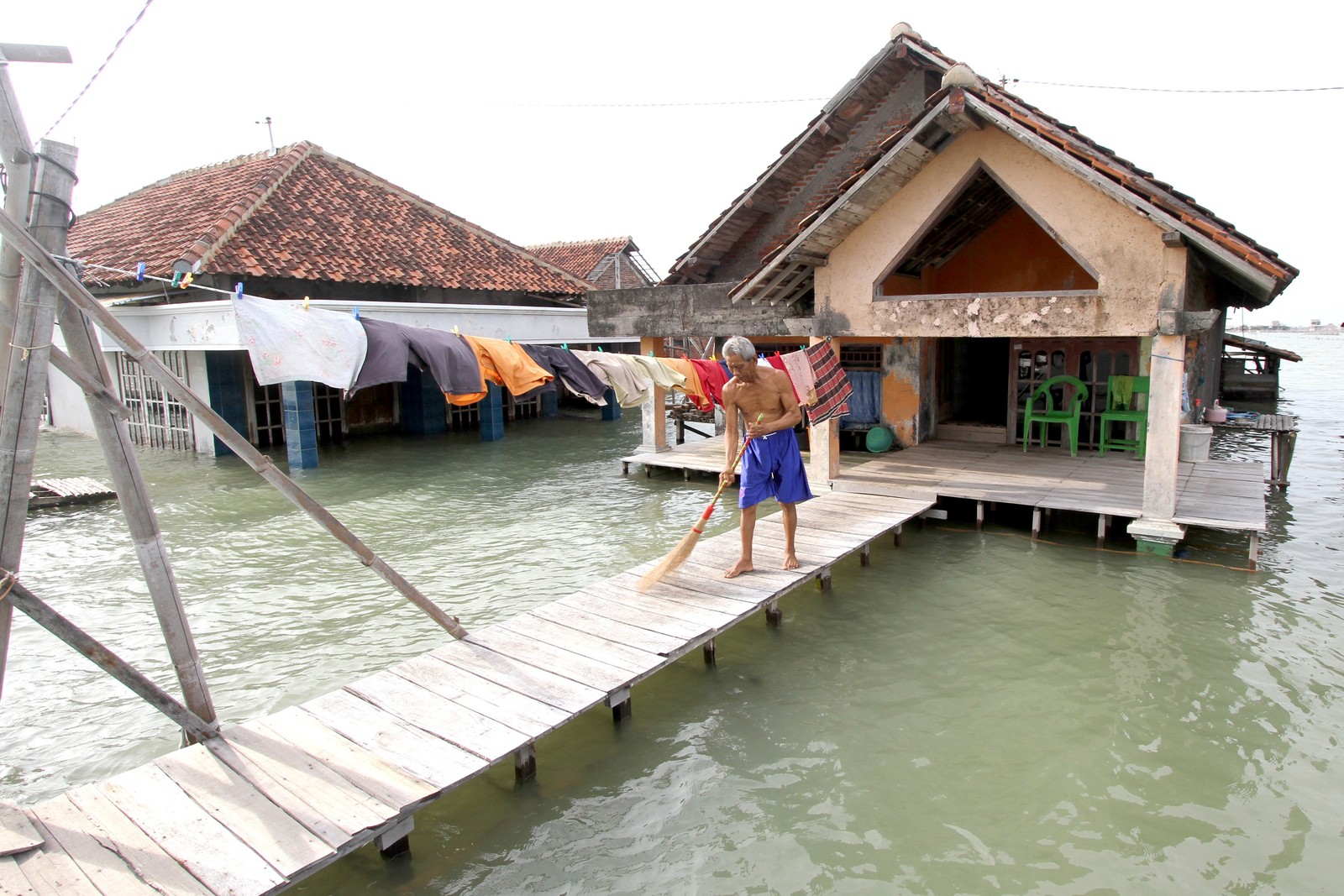 A man sweeps a boardwalk outside of a flooded house that has been surrounded by seawater.