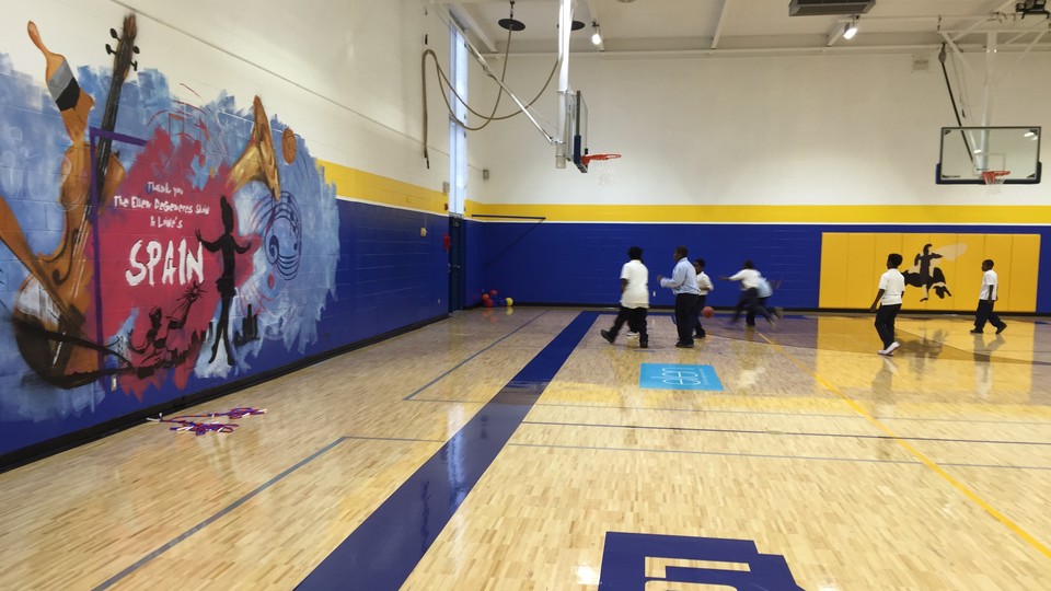 A group of children plays basketball on a brand new gymnasium floor. A mural with musical instruments and notes is pained with the school's name on the wall.