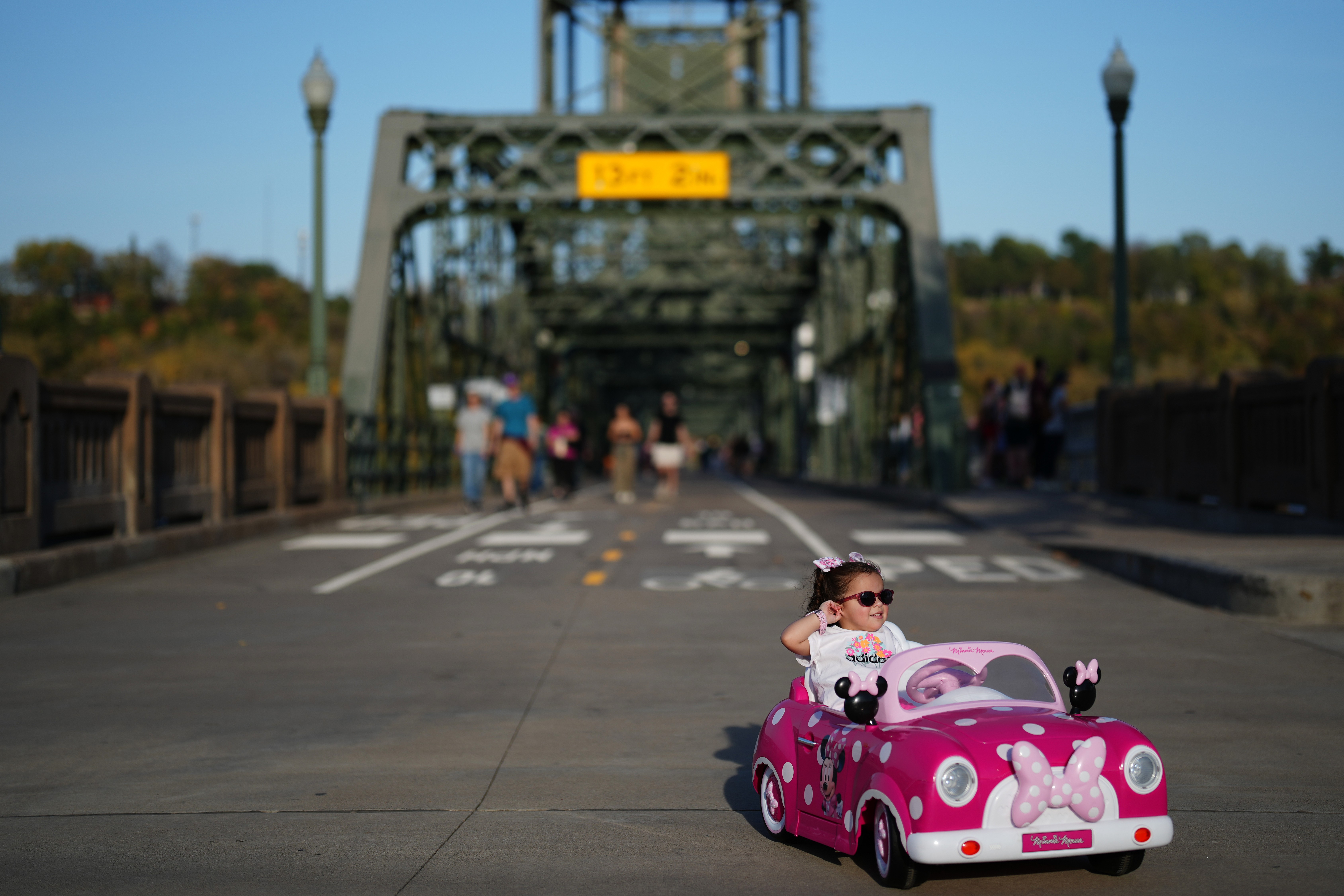 A young child drives a small pink electric car on a road open to pedestrians.
