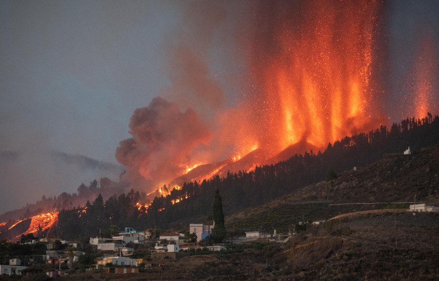 Lava and ash spew from the slope of a volcano in huge jets above several houses.