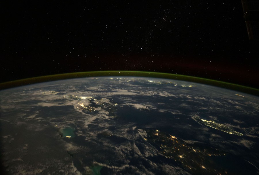 A moonlit view of the Caribbean, showing many clouds, islands, and city lights, beneath a starry sky