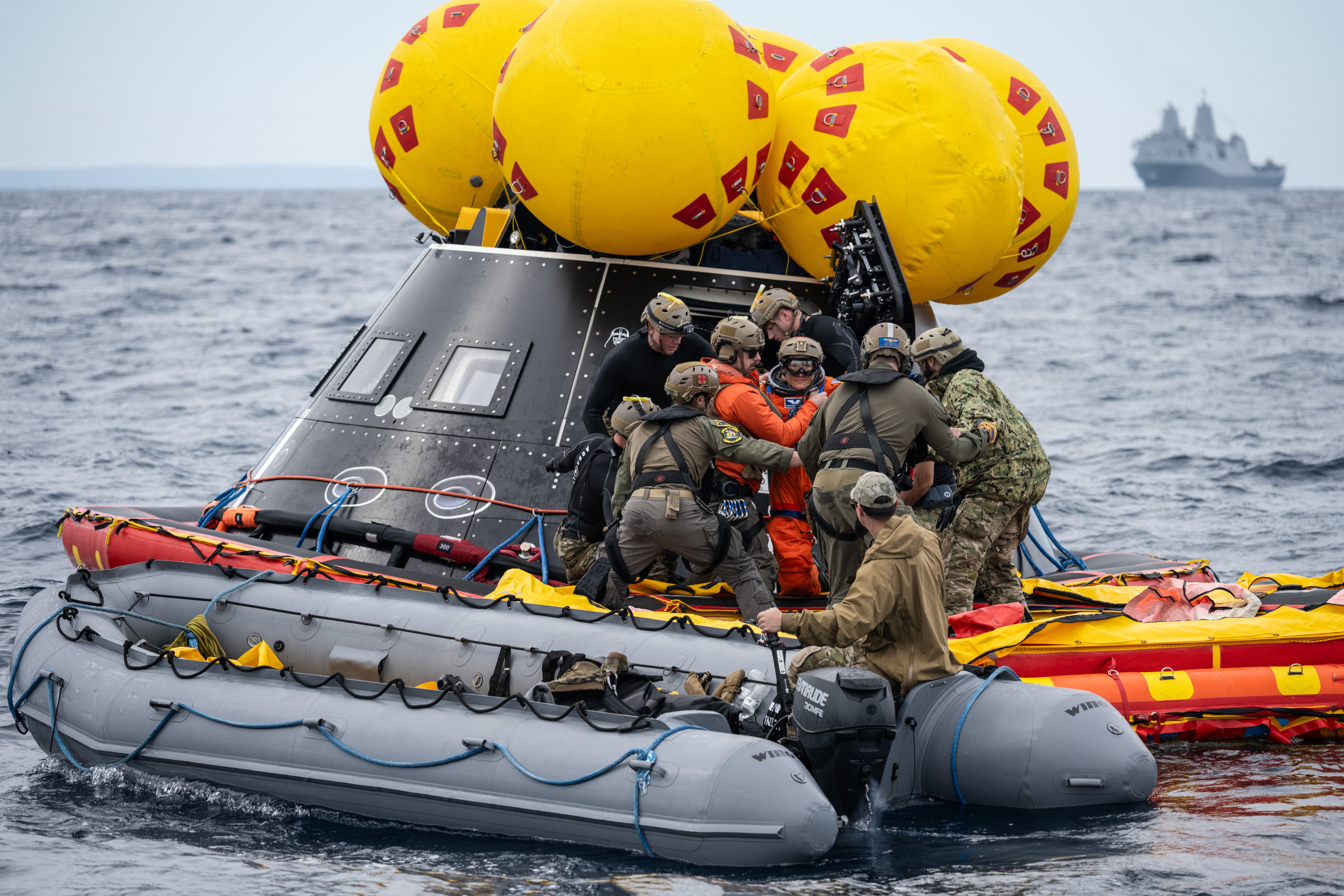 Several uniformed navy personnel work together alongside a floating spacecraft, module, helping astronauts inside get into inflatable boats.