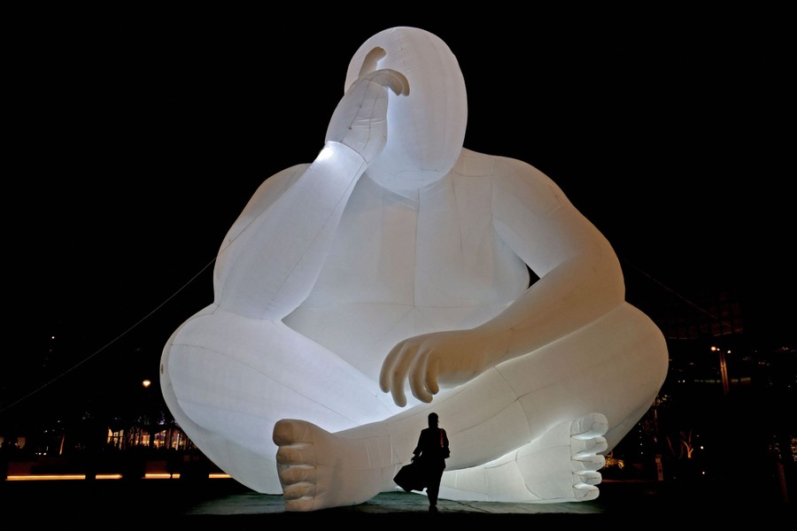 A person stands in front of an illuminated inflatable art installation in the shape of a seated human.
