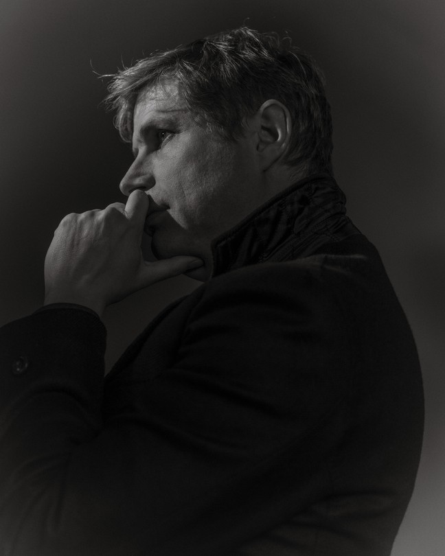 A black-and-white profile portrait of the writer Daniel Kehlmann in Berlin, his hand raised to his chin