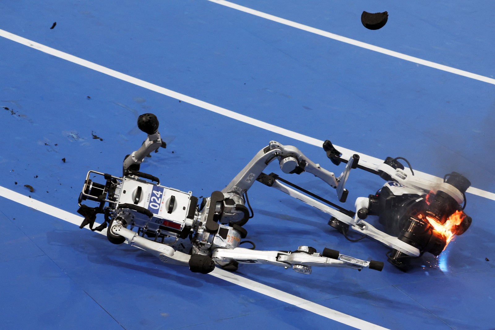 A humanoid robot falls on a track beside a burning robot competitor after a race.