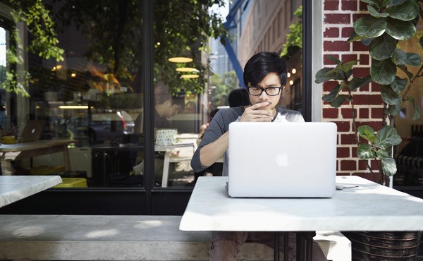 A young man looks thoughtfully at a laptop while sitting at a cafe