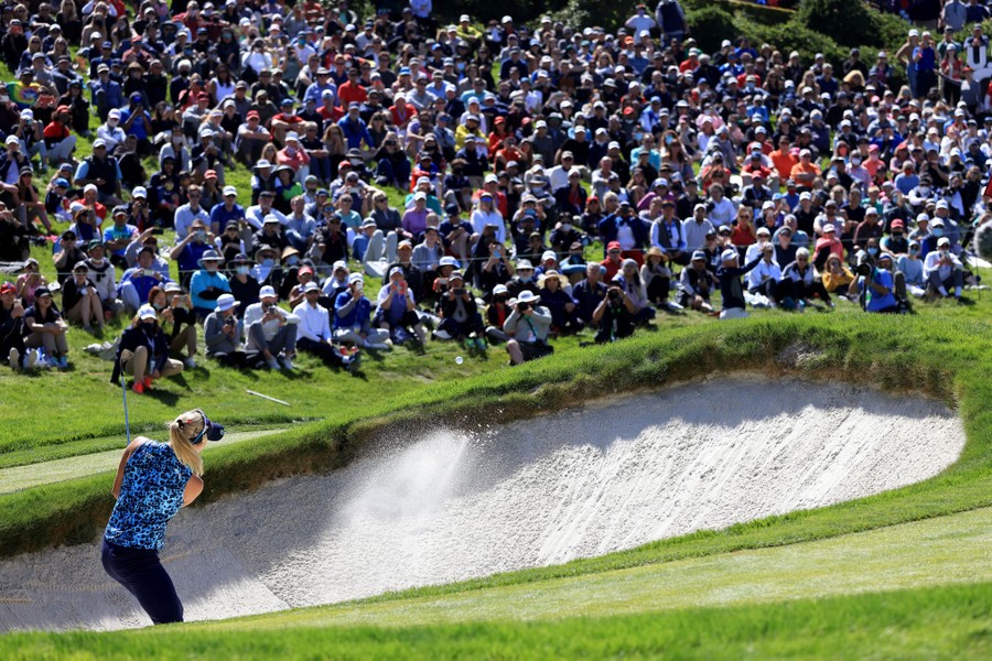 A golfer hits a ball out of a bunker as a large crowd watches.