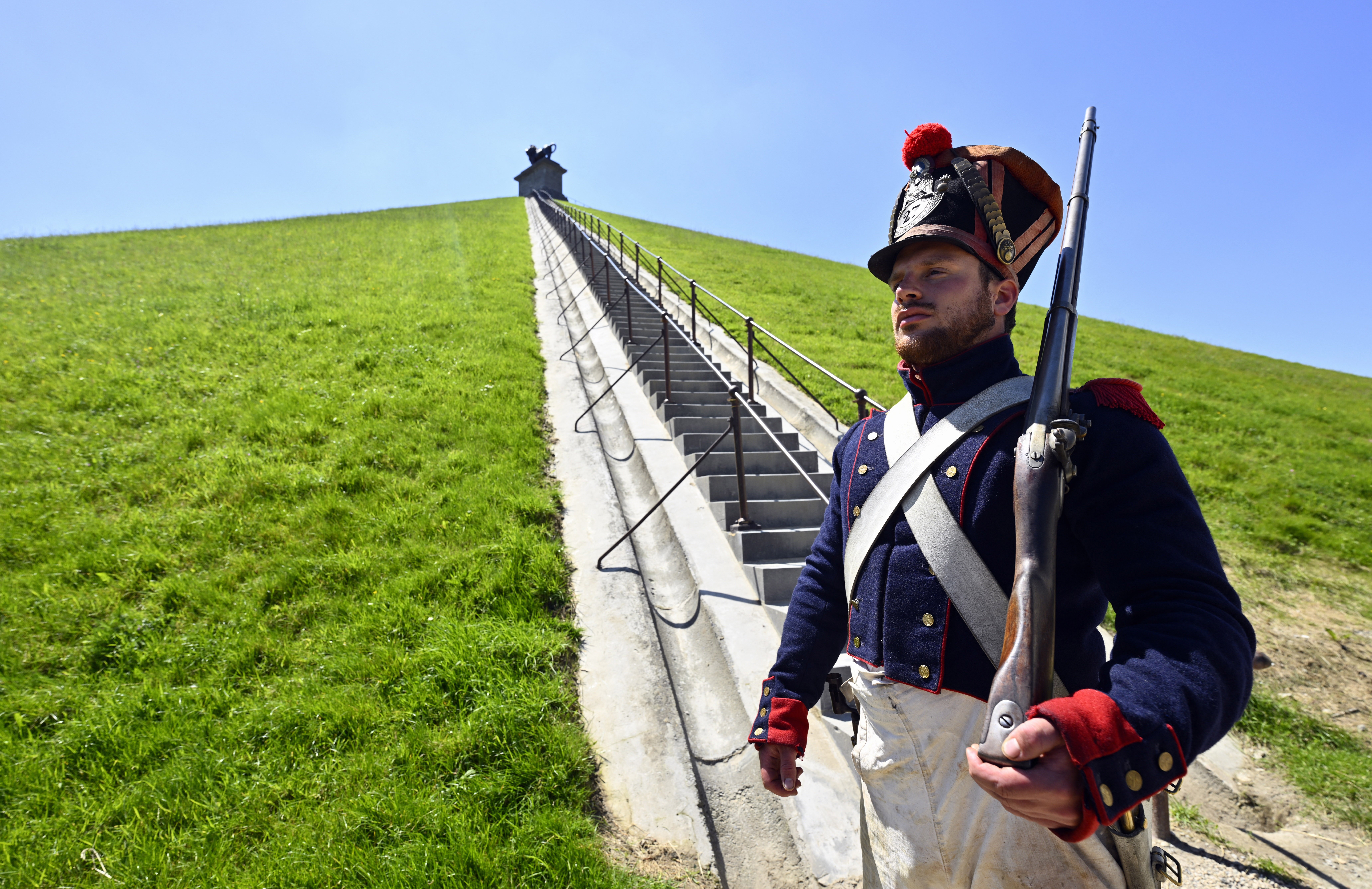 A man dressed as a member of the French Imperial Guard stands guard at the foot of a military monument at the top of a tall mound.